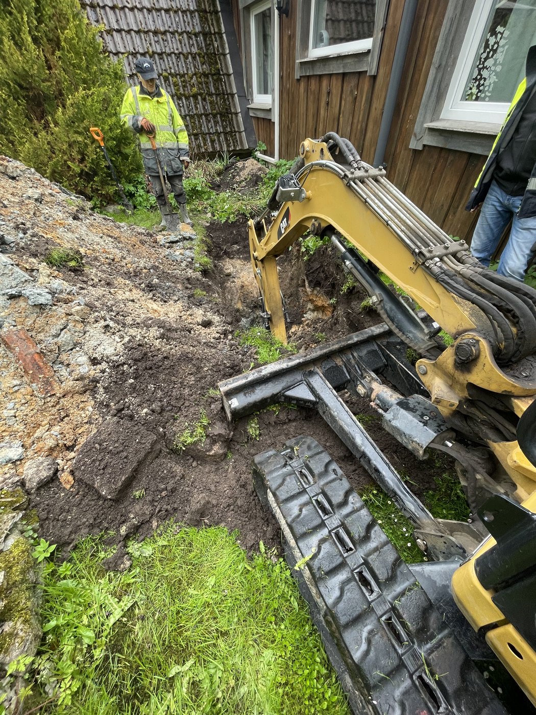 Åpen dreneringsgrøft langs husvegg med gravemaskin